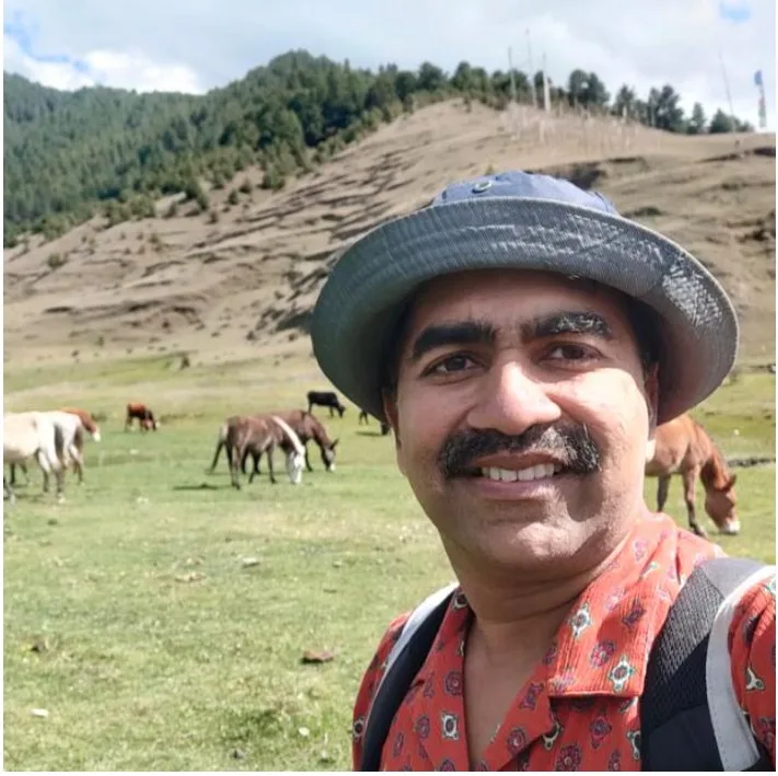 Smiling trekker with backpack in a mountain meadow with grazing horses and forested hills in the background