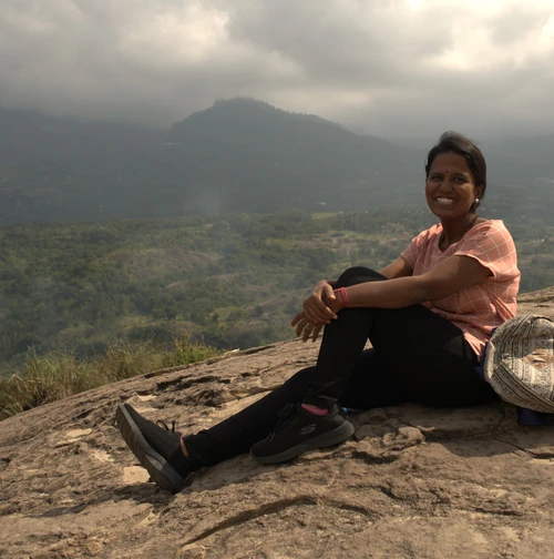 Woman trekker sitting on a rocky hill viewpoint overlooking green valleys and cloudy mountain scenery